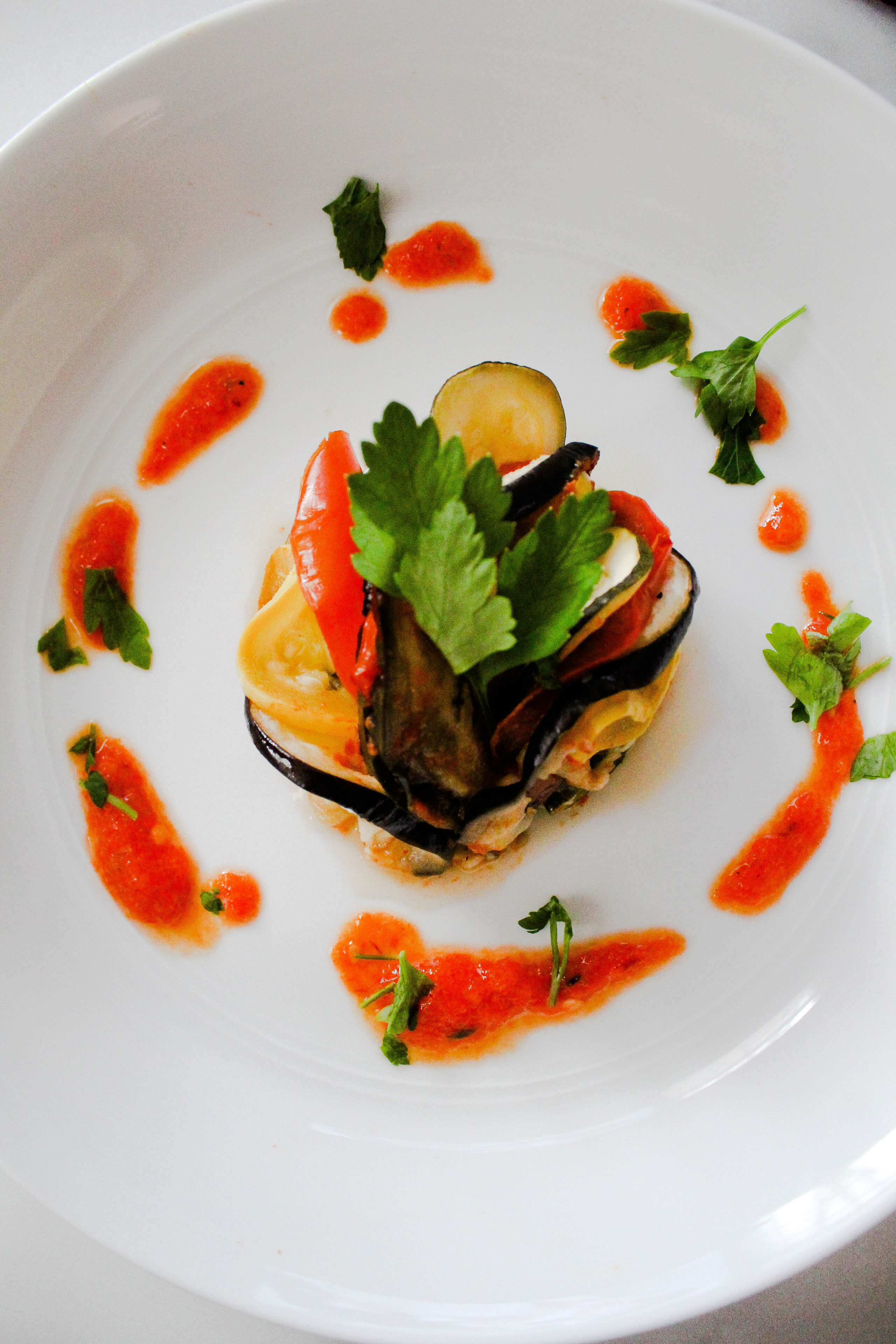 Ratatouille on a white plate surrounded by sauce and parsley.