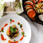 Ratatouille on a white plate surrounded by sauce and parsley sitting next to a cast iron pan with ratatouille.
