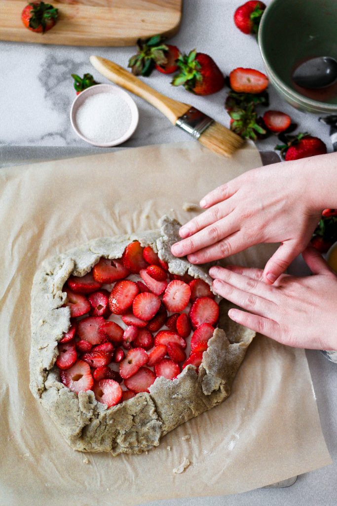 Hands folding a galette dough that is filled with strawberries.