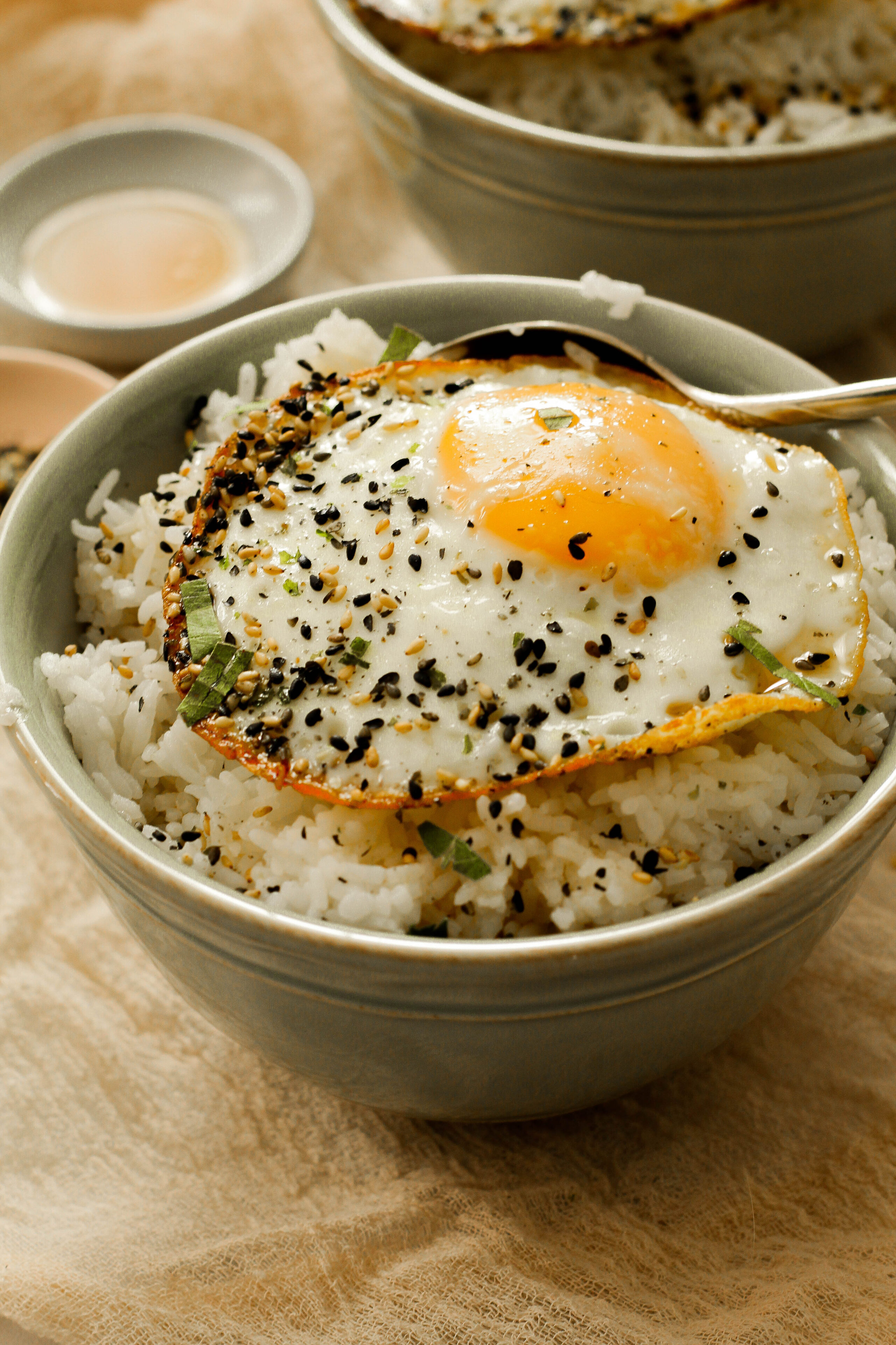 A green bowl sitting upon a biege cloth filled with rice covered in sesame sauce, a fried egg, and furikake.