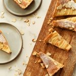 Sliced pieces of tart sitting on a cutting board as well as on two plates. Sliced almonds garnish them along with powdered sugar.