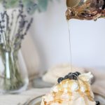 A mini beige pavlova sitting on a ceramic plate being topped with whipped cream. Syrup is being poured over it.