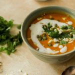 A green bowl filled with butternut squash bisque topped with cilantro, pine nuts, pumpkin seeds, and feta.
