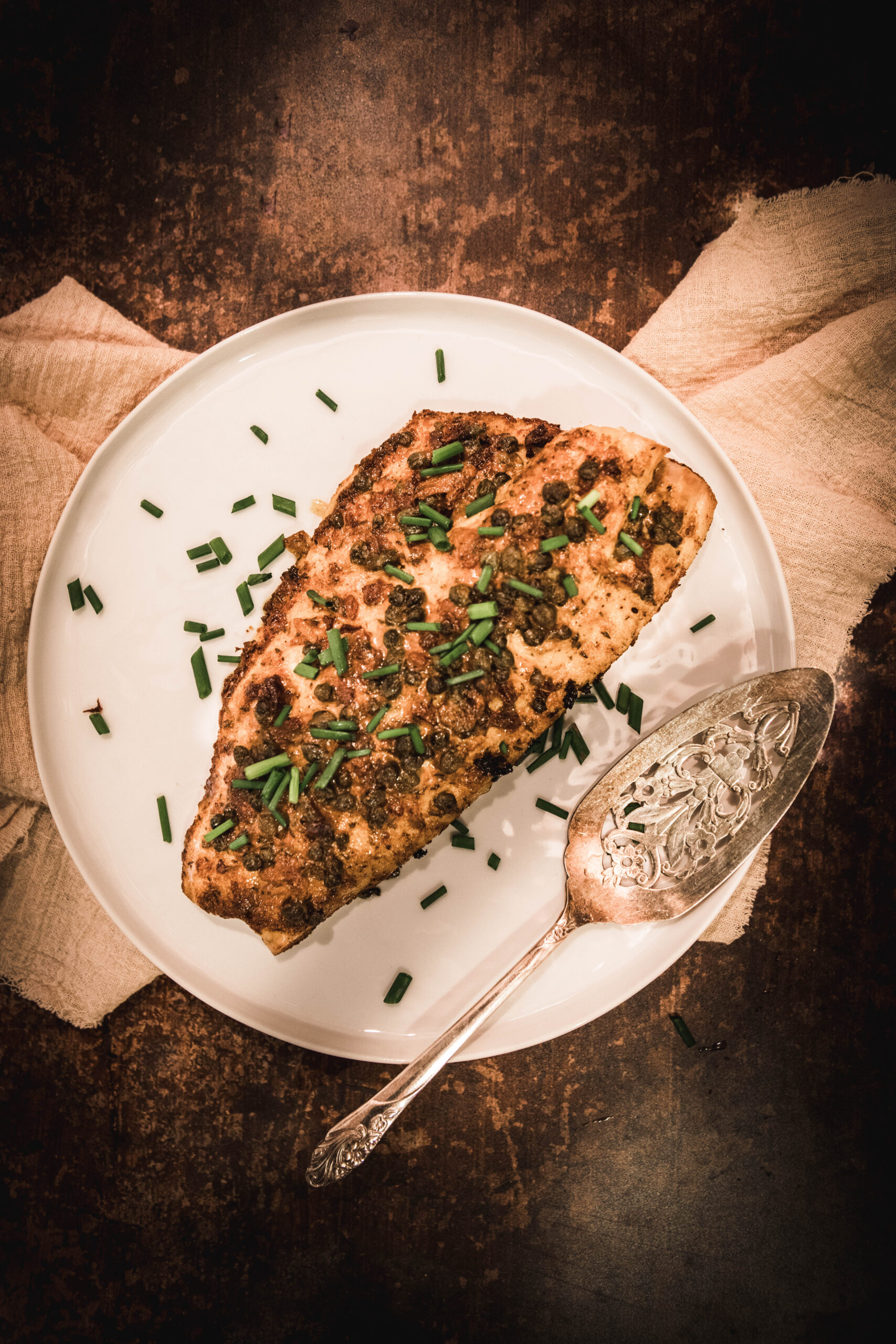 baked rockfish with chives on top on a white plate with a silver serving utensil next to it